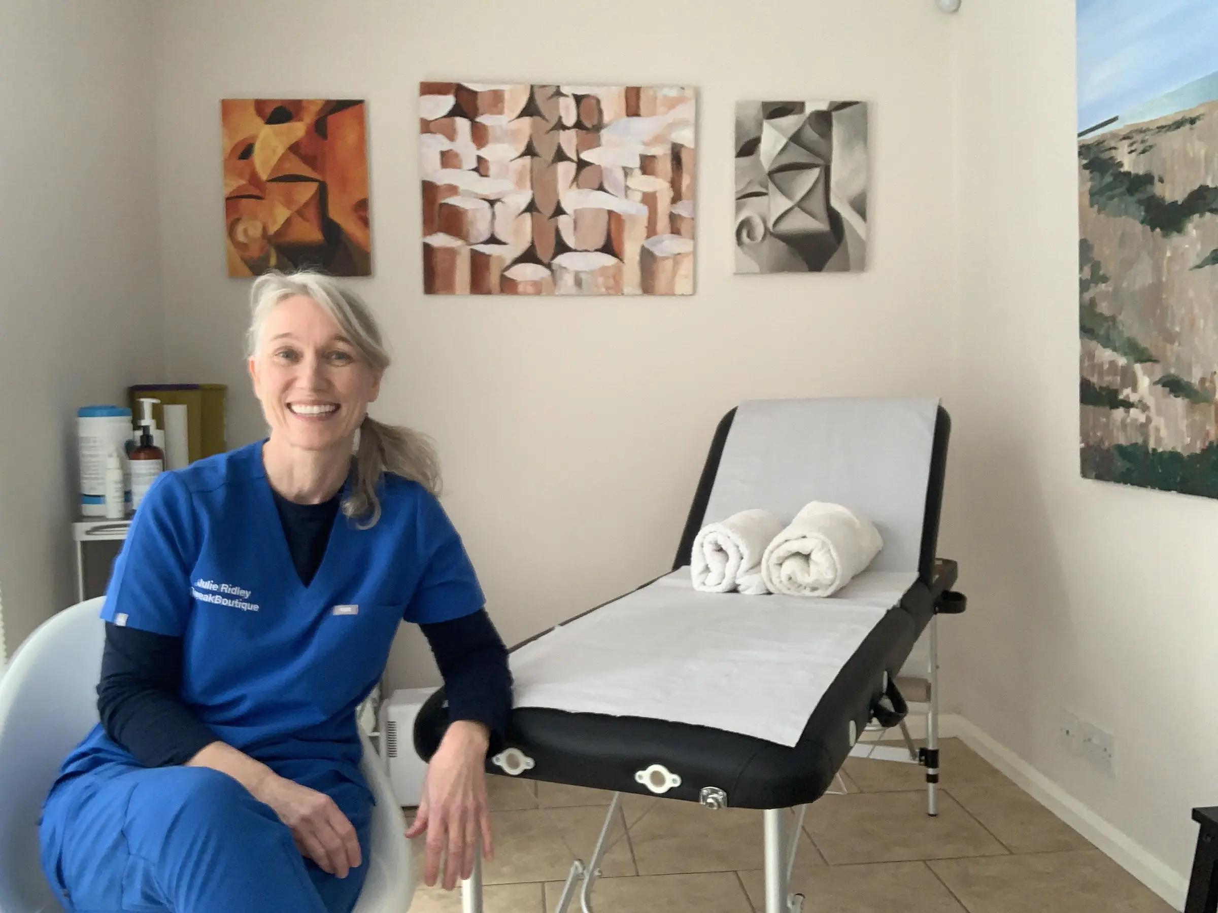 Dr Julie Ridley in blue scrubs, seated beside the treatment couch in her clinic